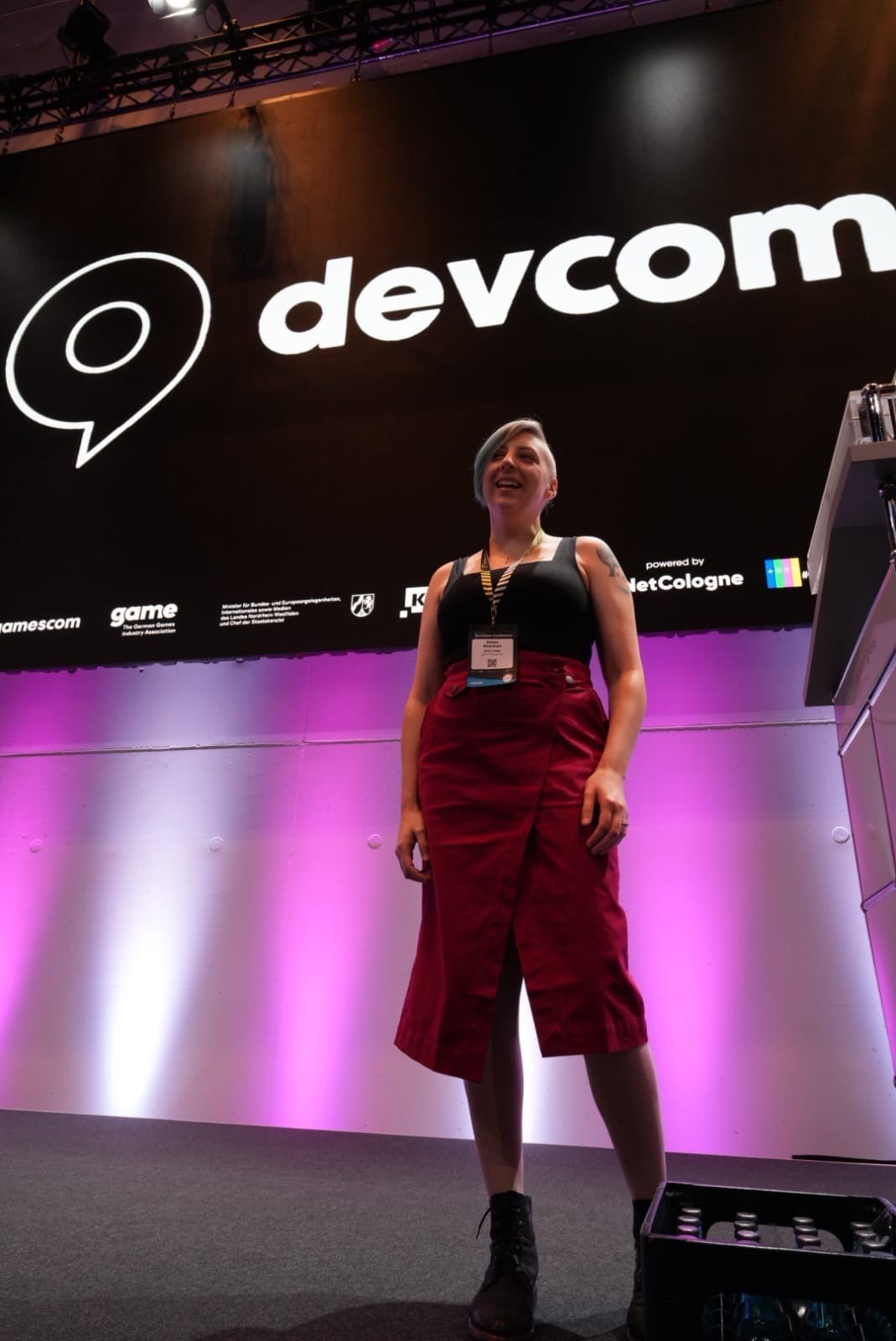 Kelsey Beachum em pé no palco da devcom, sorrindo. Ela tem cabelo curto e assimétrico em tom acinzentado azulado, usa regata preta, calça vinho, botas pretas, crachá e colares, com tatuagens visíveis nos braços. Ao fundo há um grande painel preto com o logotipo branco da devcom e iluminação de palco em tons brancos e rosados.